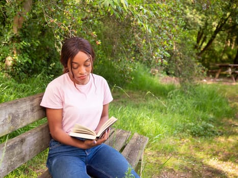 A woman is seated on a bench, engrossed in reading a book in a public park on a sunny day.