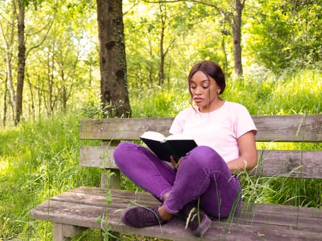 Young black woman sitting on a park bench reading a book.