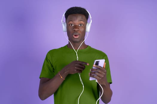 Young black man amazed while listening to music with headphones and a mobile phone on a violet background, with copyspace.