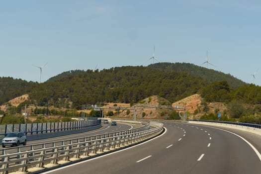 A car drives down a highway running alongside a towering mountain, under a clear sky.