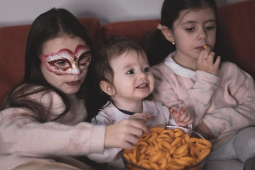 Three beautiful caucasian girls sisters are sitting on the sofa,eating corn rings and emotionally watching a movie in the room in the evening,close-up side view.The concept of family watching movies.