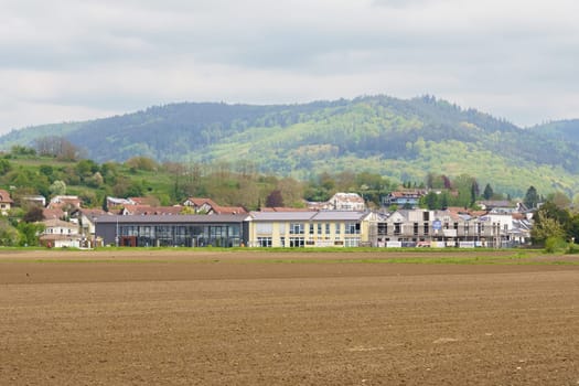 Friesenheim, Germany - April 29, 2023: A view of a rural village in the foothills of a mountain range. The buildings are surrounded by a large, plowed field. The trees are green, indicating spring.