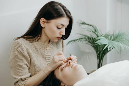 Portrait of one young beautiful Caucasian brunette cosmetologist who carefully extends artificial eyelashes with two tweezers on the left of a female client lying on a cosmetology table in the office on a spring day, close-up side view. Eyelash extension concept, beauty industry.