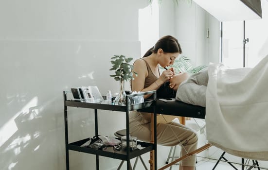 Portrait of one young beautiful Caucasian girl cosmetologist who carefully glues artificial eyelashes on the right eye with the help of two tweezers to a female client lying on a cosmetology table in her home office near the window on a spring day, close-up side view. Concept of eyelash extensions, beauty industry.