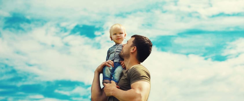 Happy strong father holding his son child on shoulder, dad with baby walking outdoors on blue sky background with clouds
