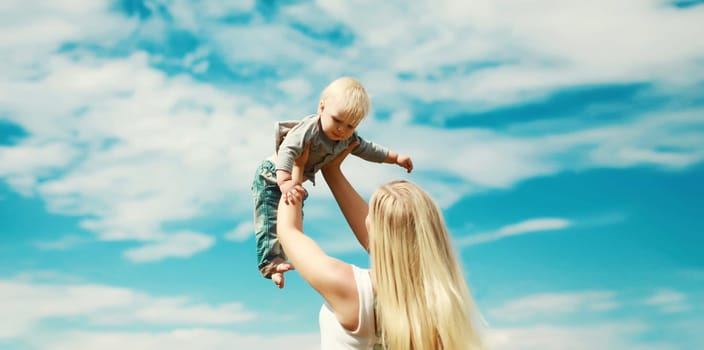 Happy young mother holding her baby raising up, joyful mom and little son child on blue sky background on a sunny summer day