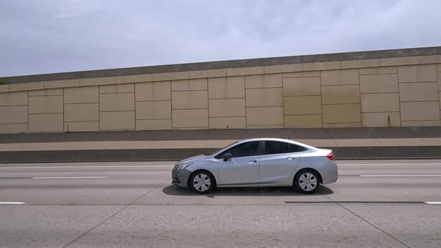 Denver, Colorado, USA-June 11, 2024-Slow motion-A scene from I-25 Highway in Denver, Colorado, capturing vehicles in motion, including a white SUV and a silver sedan. The backdrop features office buildings and lush green trees under a cloudy sky, illustrating typical highway traffic in this bustling city.