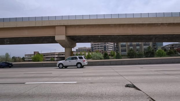 Denver, Colorado, USA-June 11, 2024-Slow motion-A scene from I-25 Highway in Denver, Colorado, capturing vehicles in motion, including a white SUV and a silver sedan. The backdrop features office buildings and lush green trees under a cloudy sky, illustrating typical highway traffic in this bustling city.