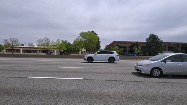 Denver, Colorado, USA-June 11, 2024-Slow motion-A scene from I-25 Highway in Denver, Colorado, capturing vehicles in motion, including a white SUV and a silver sedan. The backdrop features office buildings and lush green trees under a cloudy sky, illustrating typical highway traffic in this bustling city.