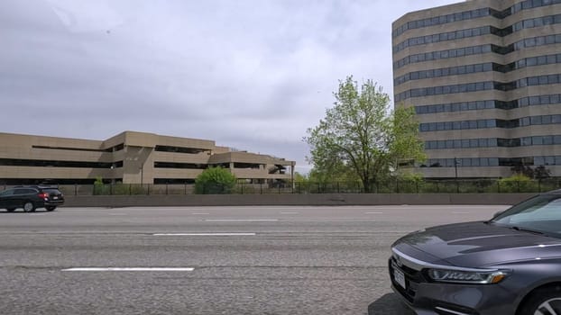 Denver, Colorado, USA-June 11, 2024-Slow motion-A scene from I-25 Highway in Denver, Colorado, capturing vehicles in motion, including a white SUV and a silver sedan. The backdrop features office buildings and lush green trees under a cloudy sky, illustrating typical highway traffic in this bustling city.