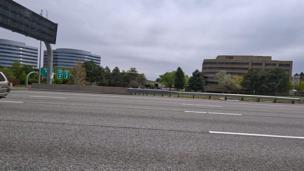 Denver, Colorado, USA-June 11, 2024-Slow motion-A scene from I-25 Highway in Denver, Colorado, capturing vehicles in motion, including a white SUV and a silver sedan. The backdrop features office buildings and lush green trees under a cloudy sky, illustrating typical highway traffic in this bustling city.