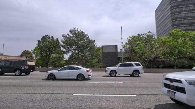 Denver, Colorado, USA-June 11, 2024-Slow motion-A scene from I-25 Highway in Denver, Colorado, capturing vehicles in motion, including a white SUV and a silver sedan. The backdrop features office buildings and lush green trees under a cloudy sky, illustrating typical highway traffic in this bustling city.