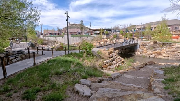 Castle Rock, Colorado, USA-June 12, 2024-Slow motion-A picturesque view of Downtown Castle Rock, Colorado, featuring a serene river flowing through the urban landscape. The scene includes a charming bridge, lush greenery, and modern buildings in the background under a bright blue sky with scattered clouds. The blend of nature and urban development highlights the unique character of this vibrant community.