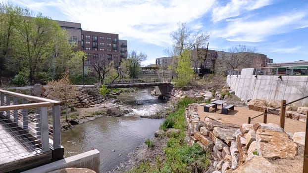 Castle Rock, Colorado, USA-June 12, 2024-Slow motion-A picturesque view of Downtown Castle Rock, Colorado, featuring a serene river flowing through the urban landscape. The scene includes a charming bridge, lush greenery, and modern buildings in the background under a bright blue sky with scattered clouds. The blend of nature and urban development highlights the unique character of this vibrant community.