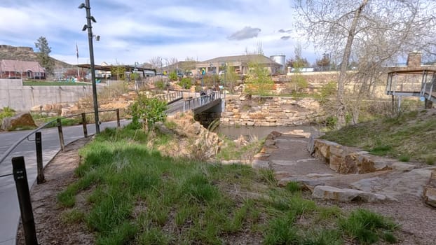 Castle Rock, Colorado, USA-June 12, 2024-Slow motion-A picturesque view of Downtown Castle Rock, Colorado, featuring a serene river flowing through the urban landscape. The scene includes a charming bridge, lush greenery, and modern buildings in the background under a bright blue sky with scattered clouds. The blend of nature and urban development highlights the unique character of this vibrant community.