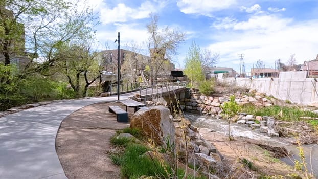 Castle Rock, Colorado, USA-June 12, 2024-Slow motion-A picturesque view of Downtown Castle Rock, Colorado, featuring a serene river flowing through the urban landscape. The scene includes a charming bridge, lush greenery, and modern buildings in the background under a bright blue sky with scattered clouds. The blend of nature and urban development highlights the unique character of this vibrant community.