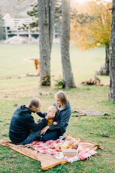 Dad touches the cheek of a little boy sitting on his mother lap on a blanket in the autumn park. High quality photo