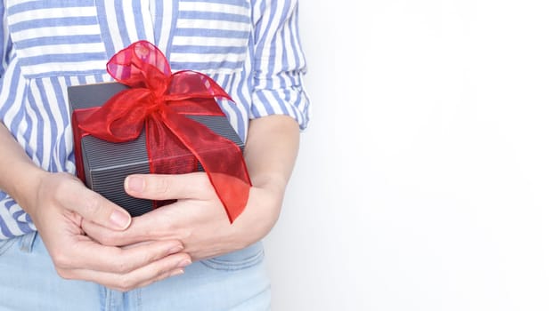 A young woman smiling while holding a beautifully wrapped gift box with a red ribbon. She is wearing a striped shirt and jeans, looking happy. The background is plain white.