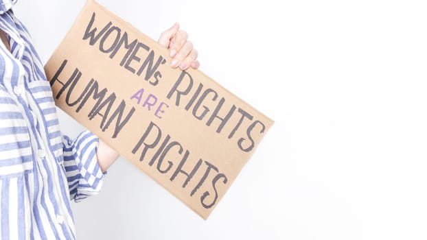 A young woman in a blue-striped shirt holds a sign that says Womens Rights are Human Rights. She looks serious, facing the camera against a white background in good lighting.