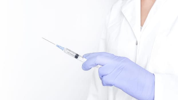 A gloved hand is holding a syringe with a needle. The syringe is filled with a clear liquid. The hand is holding the syringe in a vertical position. The needle is pointing up.