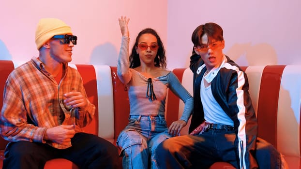 Happy girl looking at camera and moving to pop music while sitting at sofa restaurant. Hispanic woman staring at camera while dancing to freestyle beat with diverse friend in neon light. Regalement.