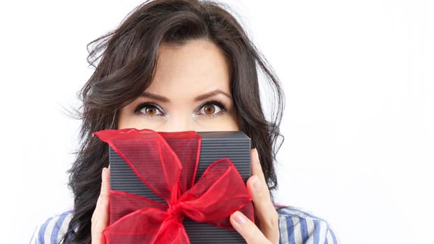 A young woman with long brown hair is holding a black gift box with a red ribbon. She is looking at the camera with a surprised expression. The woman is wearing a blue and white striped shirt.
