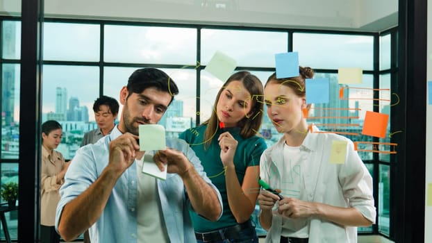 Professional business team brainstorm, sharing, thinking idea while skilled male leader write on sticky notes at glass wall. Skilled manager placed business plan on glass wall at office. Tracery
