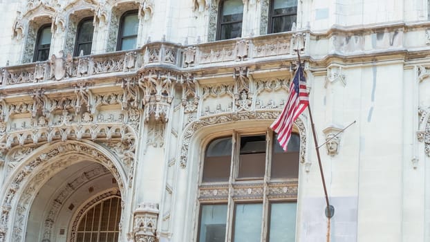 The image shows a fragment of a white vintage building with an American flag hanging from the top floor. The facade is decorated with intricate carvings and sculptures.