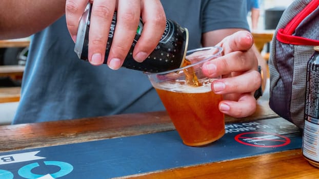A persons hand is pouring amber beer from a can into a clear plastic cup on a wooden table outdoors at a bar or restaurant. The background is blurred, and the focus is on the hand and the beer.