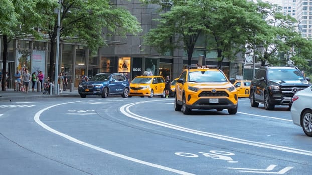 A yellow taxi cab drives through a busy city street with skyscrapers and office buildings in the background. Taxi cab is centered, background slightly blurred. Low angle shot, focused on taxi cab.