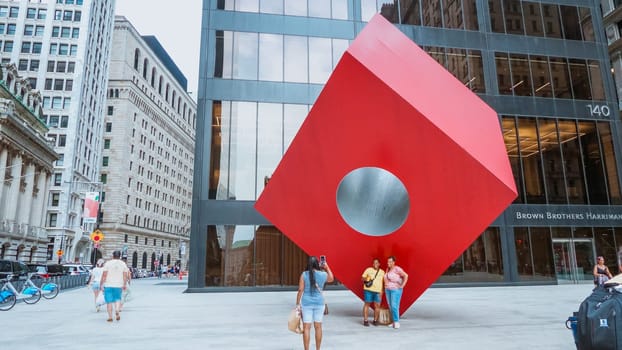 A striking red cube sculpture in front of a modern building. Its unique design with a central void catches the eye of passersby, adding an artistic touch to the urban setting.