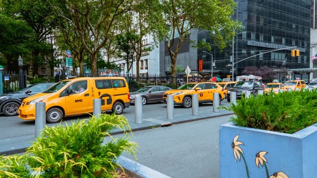 A yellow taxi cab drives through a busy city street with skyscrapers and office buildings in the background. Taxi cab is centered, background slightly blurred. Low angle shot, focused on taxi cab.