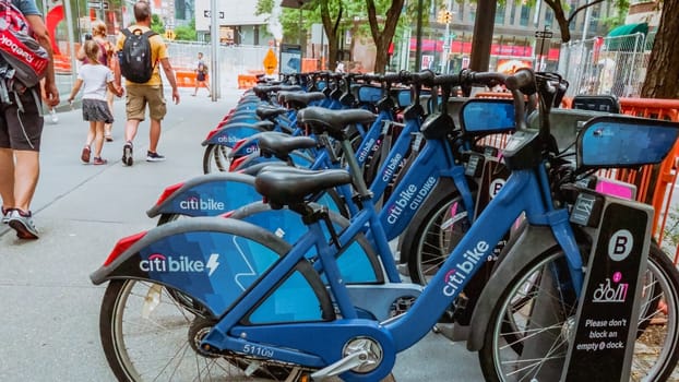 A row of vibrant blue bicycles neatly lined up on a bustling city street. Pedestrians walk by as tall buildings form a picturesque urban backdrop, creating a colorful scene in the city environment.