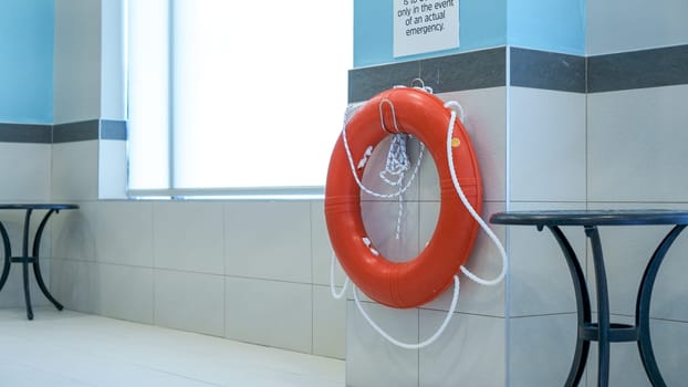 In a serene setting, a life preserver hangs on the pool wall. Lounge chairs and tables are placed nearby, creating a relaxing atmosphere by the window overlooking the pool.