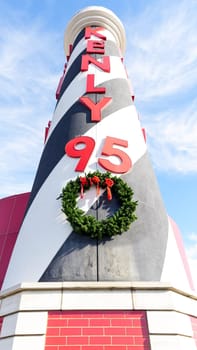 Tall black-and-white lighthouse sign with red accents in front of a historic building, under a clear blue sky with white clouds. Gas pumps and cars in parking lot by the lighthouse.