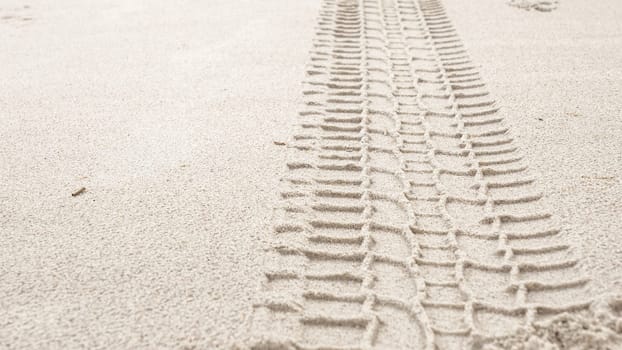 A detailed view of a wide tire track in light brown sand, likely from a heavy vehicle. The dark brown track, approximately 12 inches wide, shows human impact on natural landscapes.