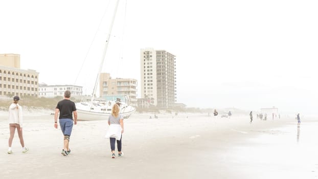 A man and woman are walking away from a beached sailboat on a foggy day. The image is taken from a low angle, making the boat look larger than life. There are buildings in the background.