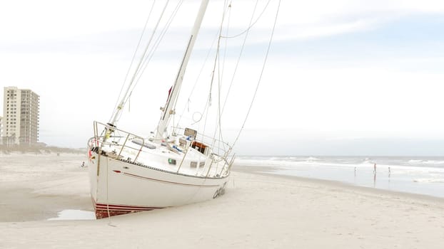 A beached yacht rests on the sandy shore after a hurricane. The damaged vessel sits askew under a gloomy sky as waves crash against the coastline, creating a scene of destruction and despair.