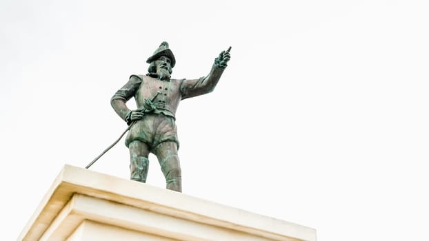 A bronze statue of Christopher Columbus stands atop a marble pedestal, pointing at the horizon with one hand while holding a map in the other. The statue is set against a solid white background.