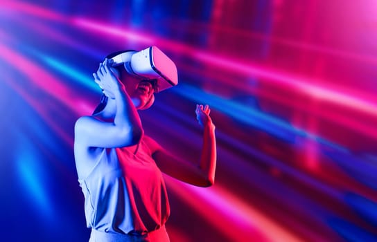 Smart female standing surrounded by neon light wearing VR headset connecting metaverse, future cyberspace community technology. Elegant woman enjoy dancing wavering body and sing song. Hallucination.