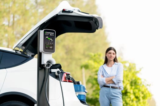 Young woman recharging battery for electric car during autumnal road trip travel EV car in autumnal forest. Eco friendly travel on vacation during autumn with electric vehicle. Exalt