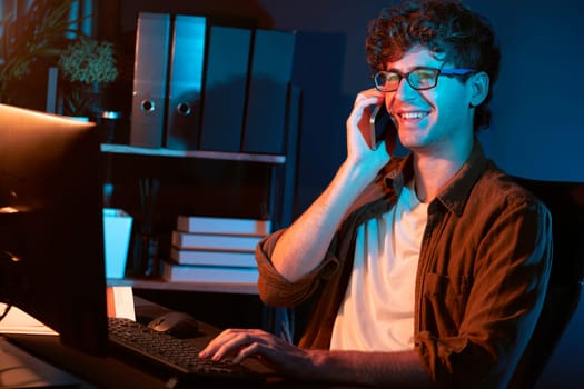 Young businessman calling on smartphone to customer or coworker, looking at computer screen of creative presentation surround work desk at neon modern office. Concept of new next project. Gusher.