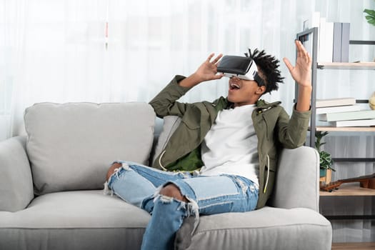Surprised African American looking VR enjoy watching by touching the air in fantastic 3D cartoon movie technology virtual reality metaverse journey new world at living room sitting sofa. Contrivance.
