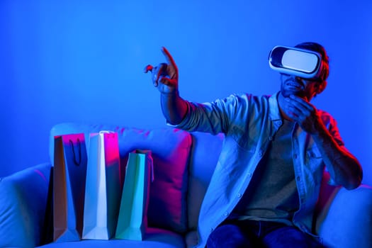 Caucasian man with vr glass posing pointing finger while sitting at sofa with shopping bags. Person selecting and doing shopping online by using VR technology and virtual goggle at home. Deviation.