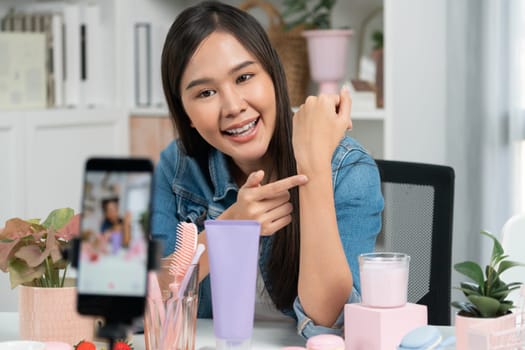 Young beautiful Asian girl reviewing moisturizing body lotion cream product to make glow skin testing on arm on social media online live recording smartphone with bestseller at modern room. Stratagem.