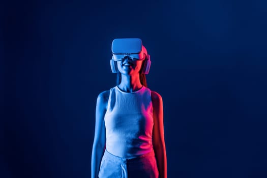 Smart female stand with surrounded by cyberpunk neon light wear VR headset connecting metaverse, futuristic cyberspace community technology. Elegant woman look generated virtual area. Hallucination.