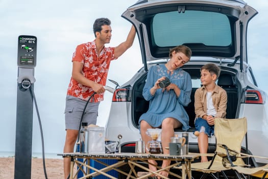 Family vacation trip traveling by the beach with electric car, happy family recharge EV car, enjoying outdoor camping coffee. Seascape travel and eco-friendly car for clean environment. Perpetual