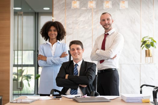 Confident diverse business team of three people, including a woman and two men, posing with arms crossed at an office desk with a laptop and paperwork, bright modern office setting