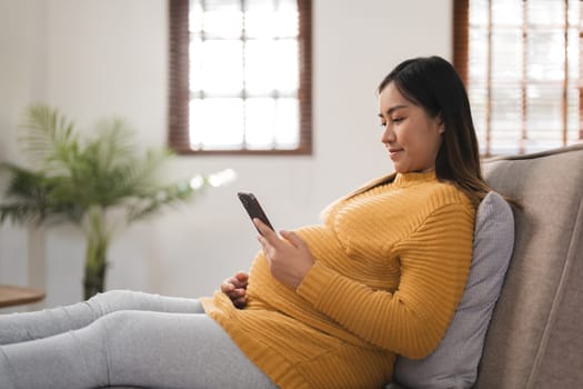 Pregnant woman sitting on a sofa in a modern living room, using a smartphone, wearing comfortable maternity clothing, with natural light and a cozy home environment.