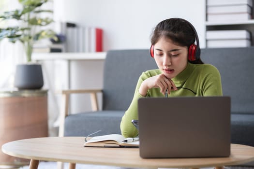 Young woman working on a laptop in a cozy living room, wearing headphones and a green sweater, focused on her tasks.